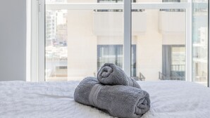 A hotel room with a large window, a bed with white bedding, and two rolled towels on the bed. - San Giljan Stylish Apartment (St. Julian's)