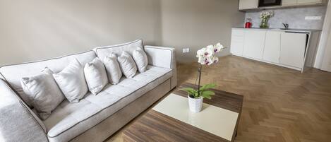 Uma sala de estar moderna com um sofá cinzento, uma mesa de centro de madeira e uma cozinha ao fundo.