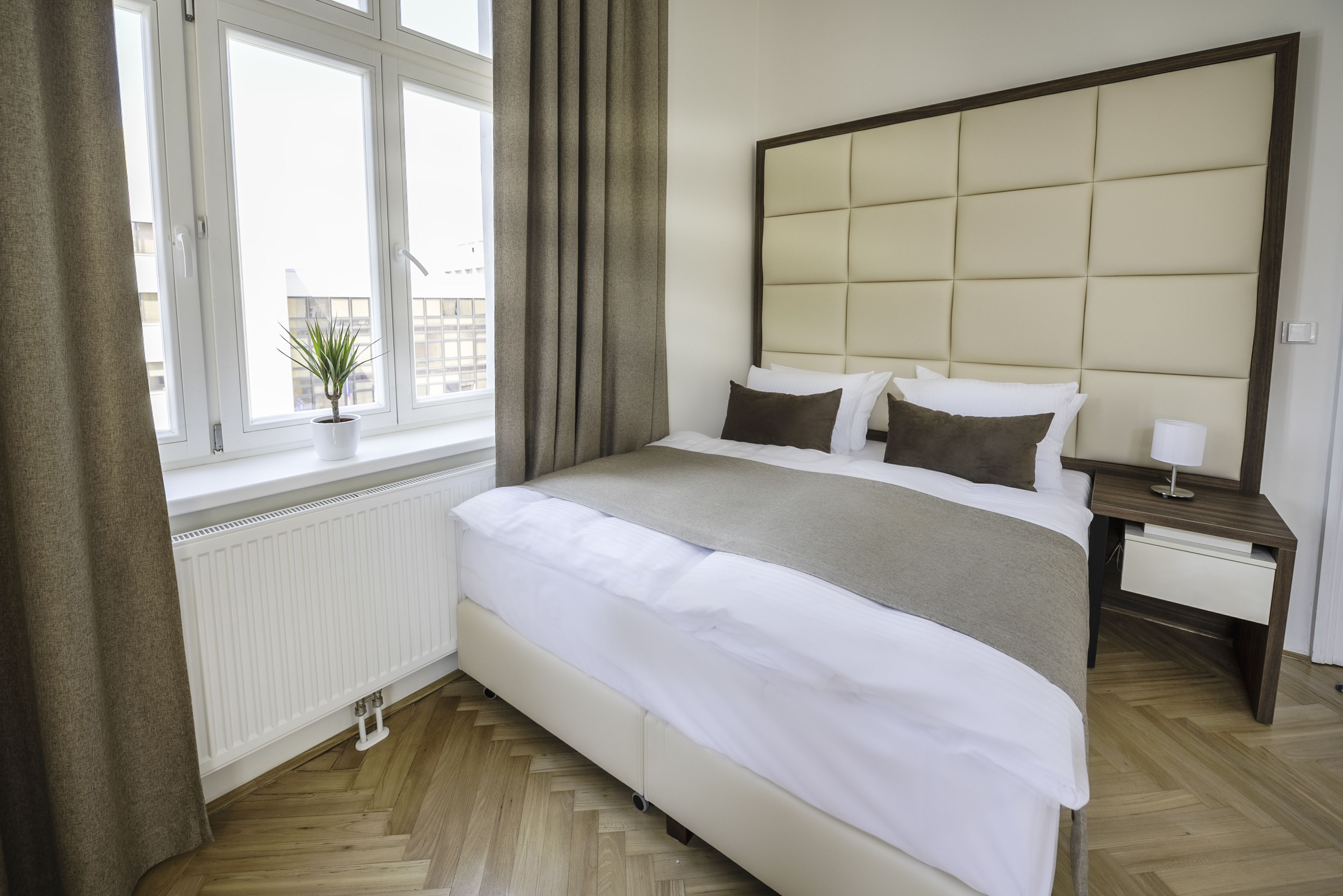 A hotel room with a large bed, a bedside table, a nightstand, a window with curtains, and a potted plant.