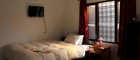 A bedroom with a bed, a nightstand with a lamp, a window with red curtains, and a wall-mounted TV.