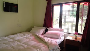 A single bed with white bedding, a bedside table with a vase of flowers, and a window with red curtains. - Gurkha Lodge (Pokhara)
