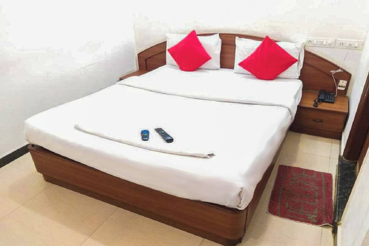 A neatly made bed with white bedding and two red pillows, a bedside table with a phone and remote controls, and a red rug on the floor.