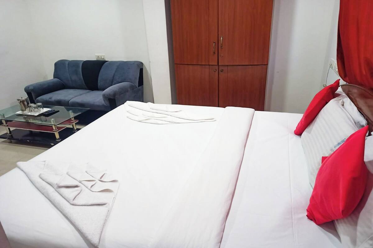 A neatly made bed with white linens and a red pillow, a glass coffee table with a book and a thermos, a blue armchair, and a wooden wardrobe.