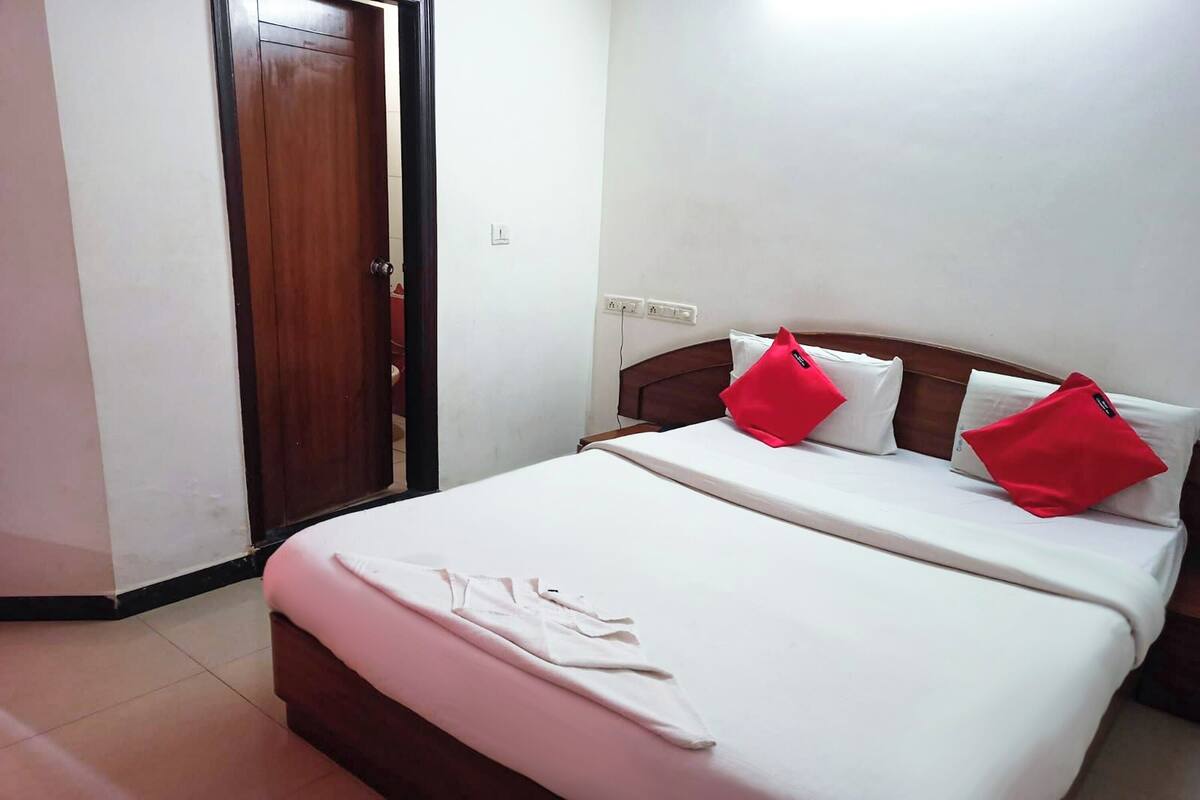 A neatly made bed with white linens and red pillows, a wooden door, and a white wall with a power outlet.