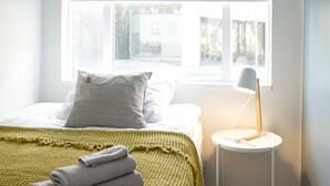 A bedroom with a bed, a window with a white blind, a small white side table with a lamp, and a green blanket on the bed. - Apartment T10c (Reykjavik)