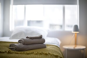 A neatly made bed with folded towels, a bedside lamp, and a window with a view of the outside.