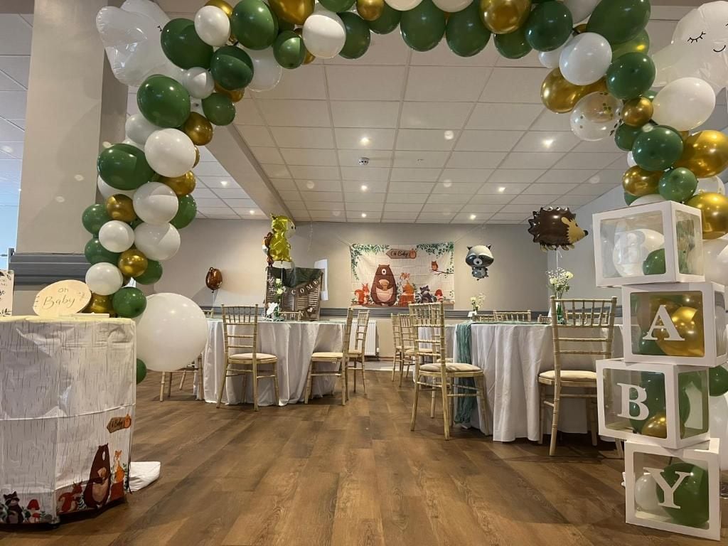 Decoración para una celebración de bienvenida al bebé, con un arco de globos, mesas, sillas y un fondo adornado con motivos infantiles.