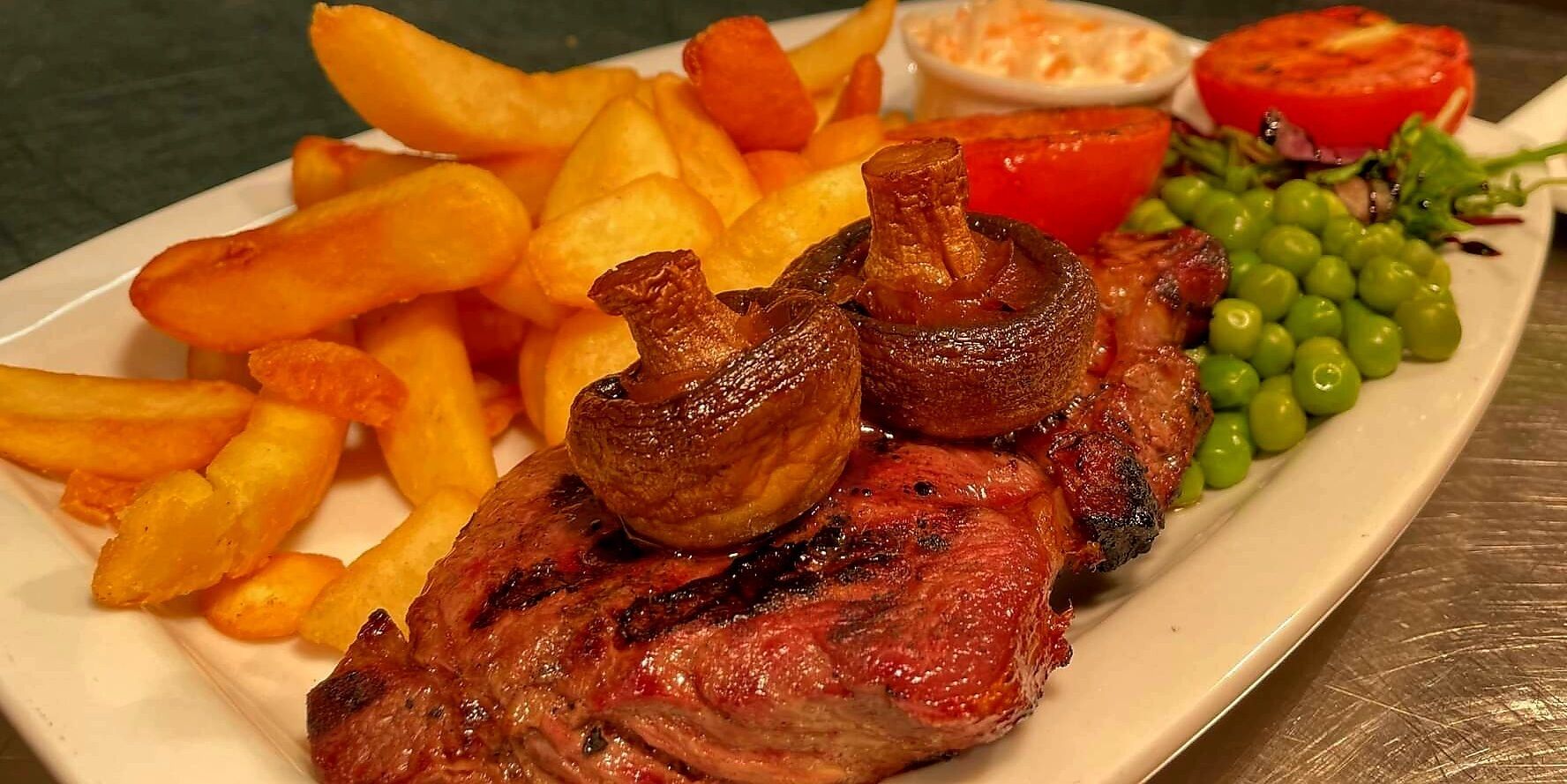 Un plato con filete a la parrilla, papas fritas, champiñones, guisantes y una rodaja de tomate.