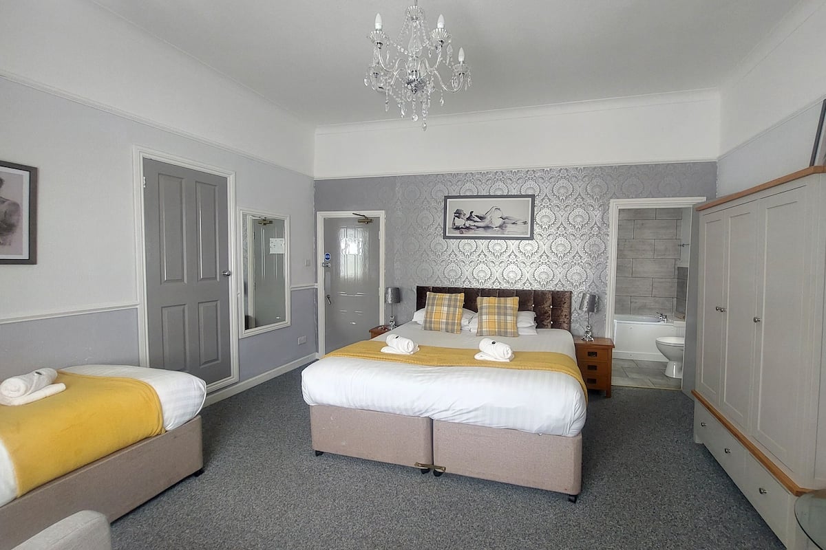 A hotel room with a large bed, two bedside tables, a wardrobe, a bathroom with a bathtub and toilet, and a chandelier.