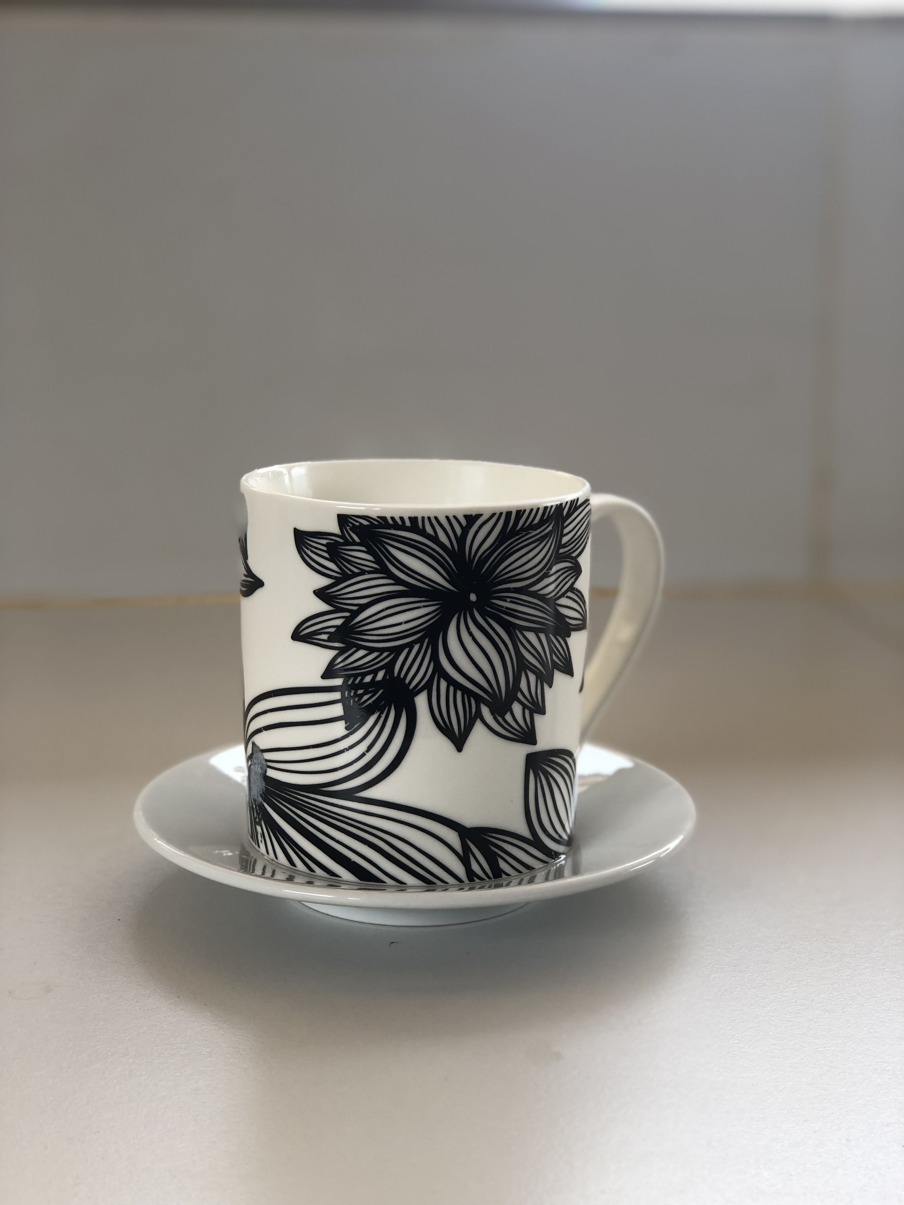 Una taza blanca con un diseño floral negro sobre un platito a juego.