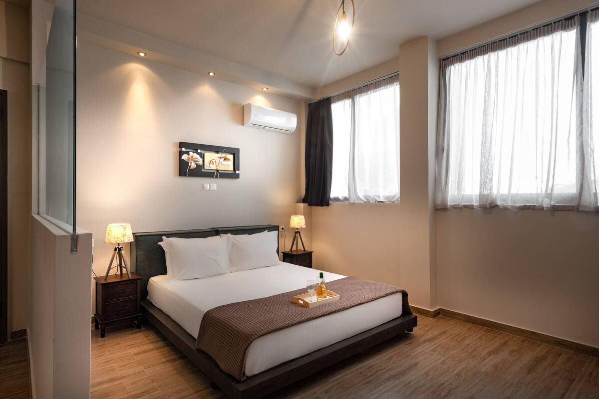 Een keurig opgemaakt bed met een dienblad met ontbijtartikelen, een wandairconditioner en een raam met dunne gordijnen.