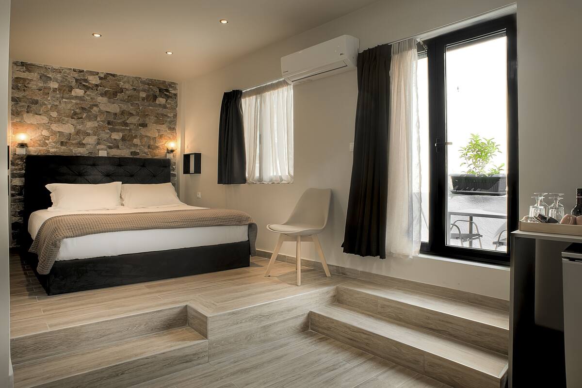 Een moderne slaapkamer met een wand van steen, een bed met een zwart hoofdeinde, een witte stoel en een groot raam met gordijnen.