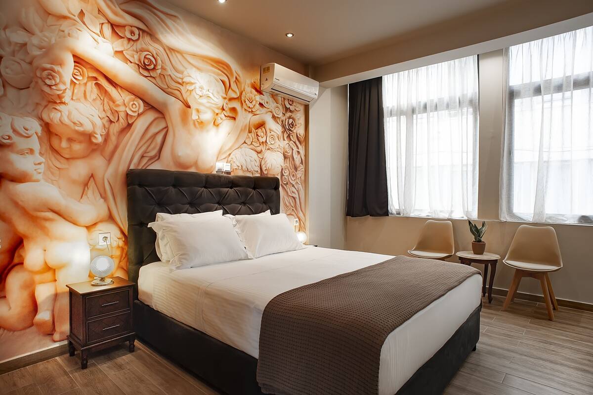 Slaapkamer met een groot bed, nachtkastje, muurschildering, raam met gordijnen en twee stoelen.