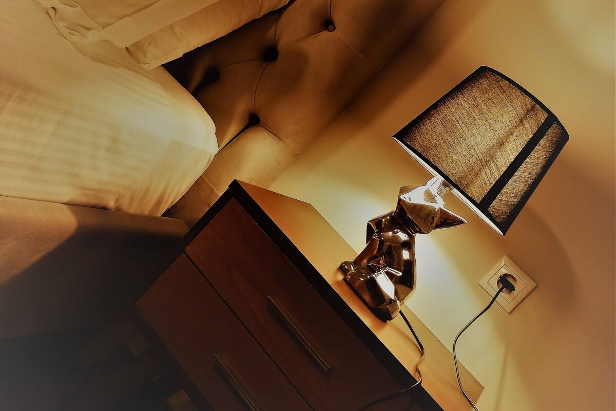 Een slaapkamer met een gestoffeerd hoofdeinde, een nachtkastje met een lamp en een houten dressoir.