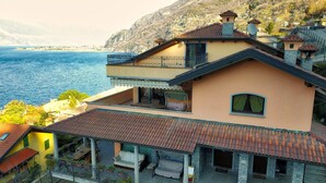 A two-story house with a balcony overlooking a body of water, a patio area with outdoor furniture, and a mountainous backdrop.