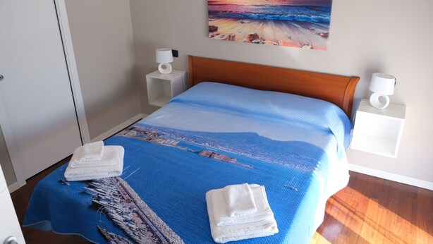 A neatly made bed with a blue bedspread and two towels, a painting of a sunset beach on the wall, and a wooden headboard. - Turist Rent Apartment Nadia (Napoli)