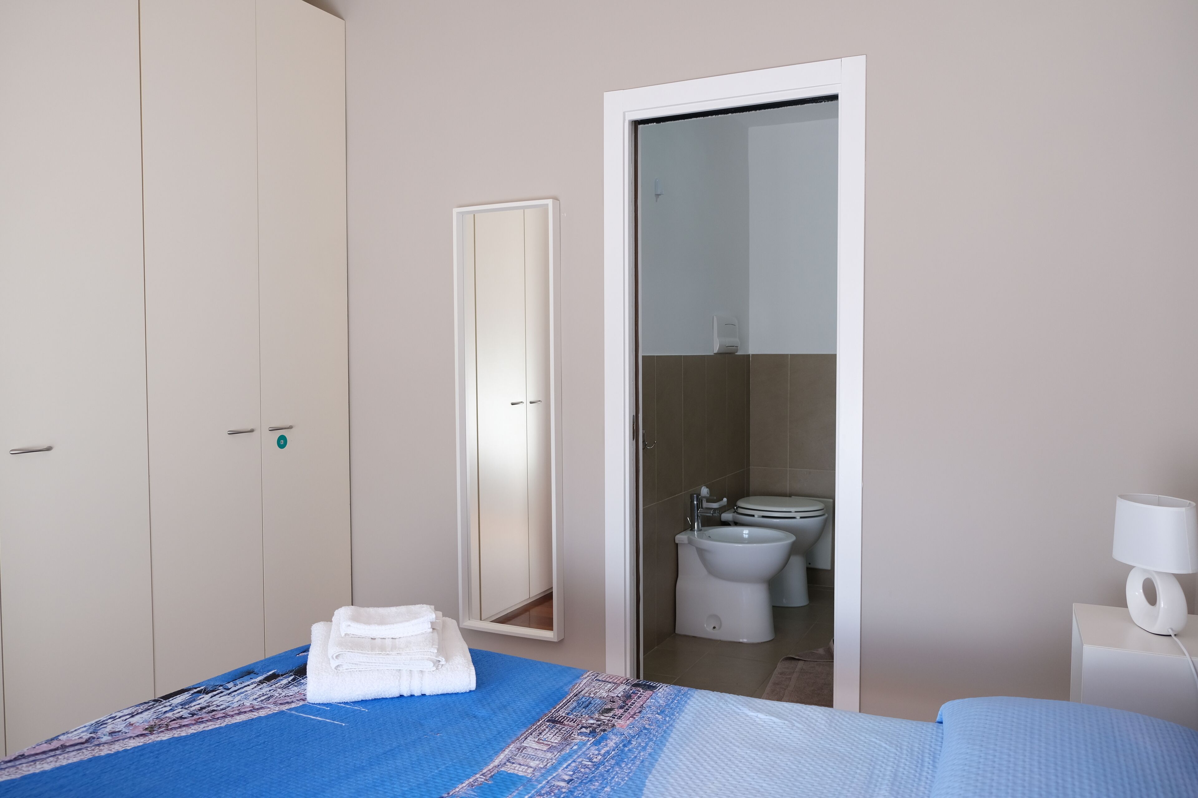 Une chambre à coucher avec un lit, une table de chevet avec une lampe, une armoire et une salle de bain visible à travers une porte ouverte.