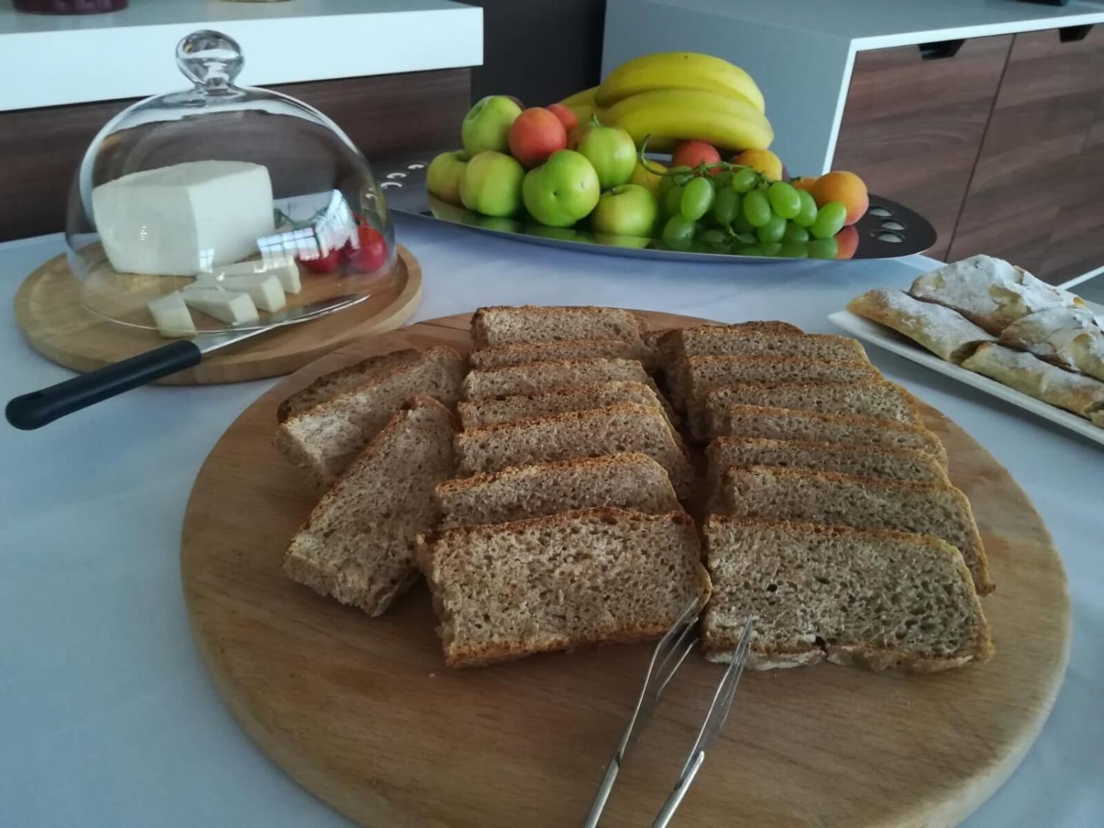 A wooden cutting board with sliced bread, a glass cheese container, a fruit bowl with apples and bananas, and a plate of pastries.