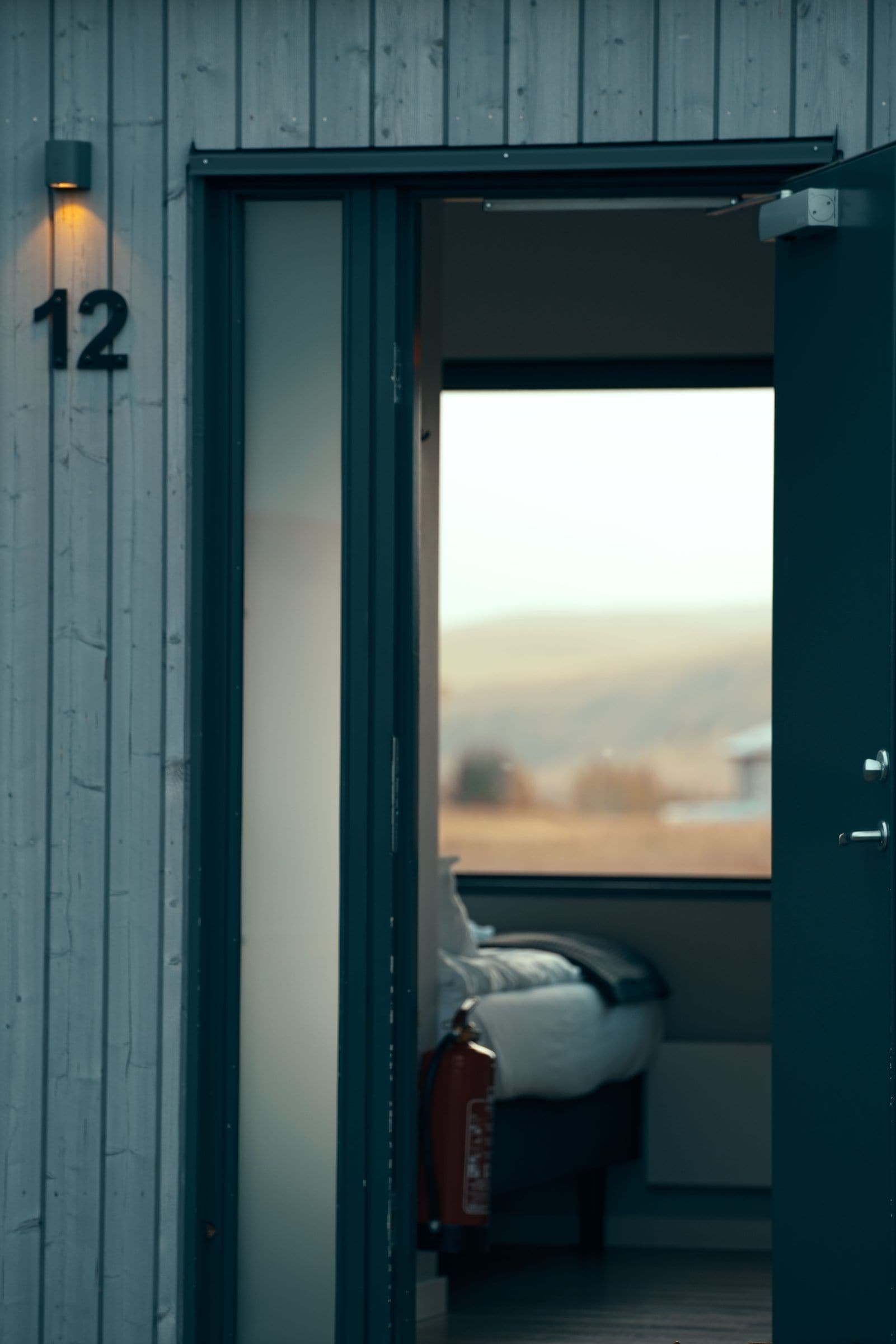 A room with a bed, a fire extinguisher, and a window with a view of the outdoors.