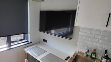 A compact kitchen with a built-in TV, a dining area with a chair, and a window with a blind.