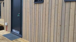 Un edificio moderno con revestimiento de madera vertical, una puerta negra y dos ventanas pequeñas.
