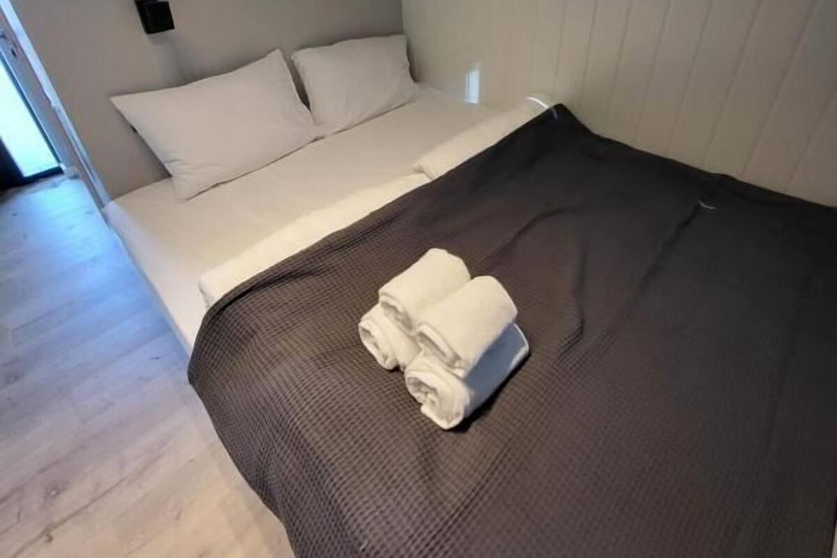 Yhden hengen sänky, jossa on tummanruskea patja, valkoiset tyynyt ja valkoinen peitto. Sängyllä on kaksi rullalle taitettua valkoista pyyhettä.