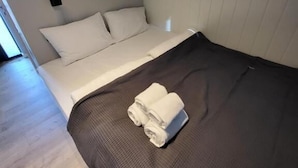 Una cama individual con colchón marrón oscuro, almohadas blancas y edredón blanco. Hay dos toallas blancas enrolladas sobre la cama.