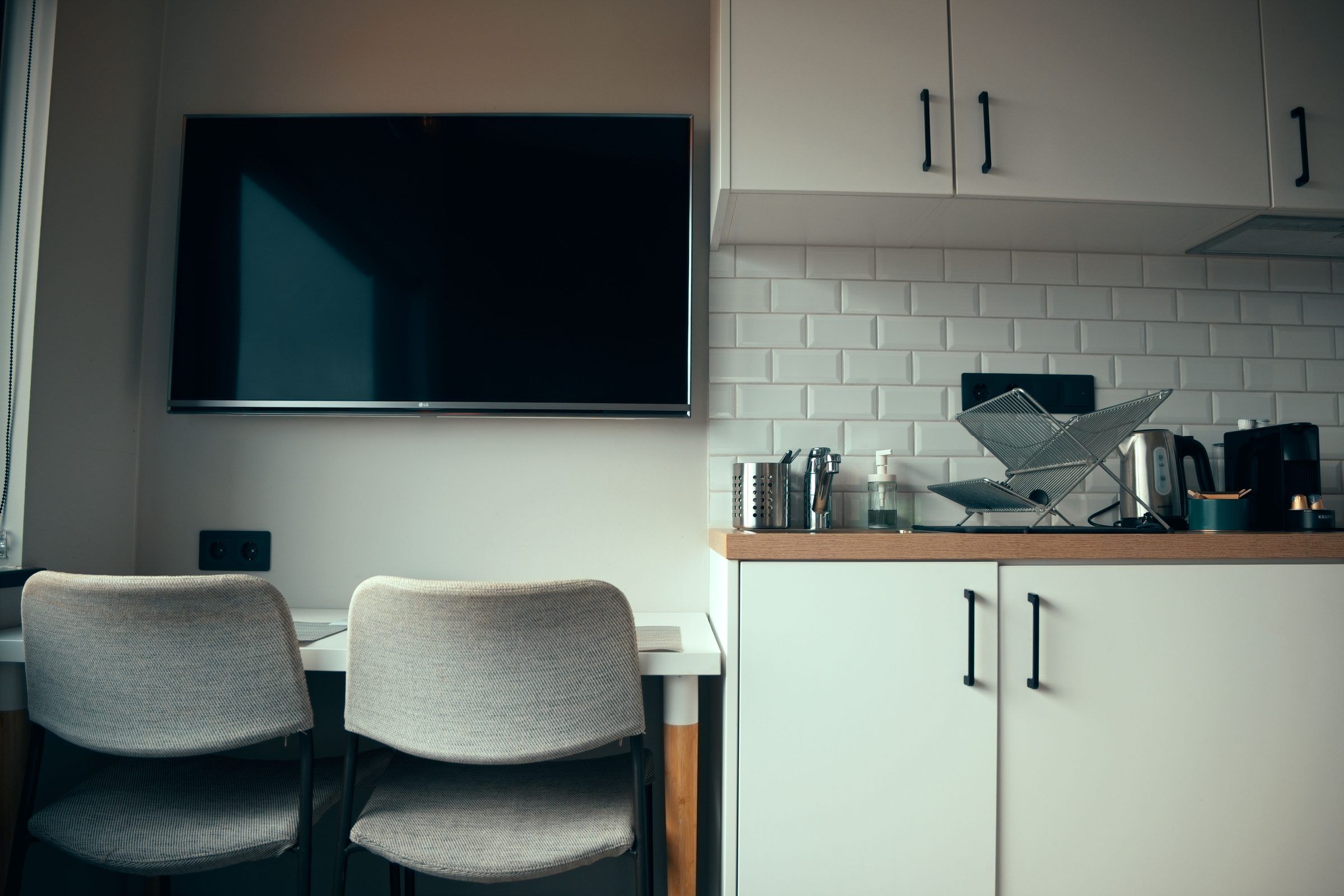 A modern kitchen with white cabinets, a large flat-screen TV, and a dining area with two chairs.
