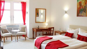 A hotel room with a bed, a desk, two chairs, a radiator, and a window with red curtains. - Biohotel Schloss Kirchberg (Kirchberg an der Jagst)