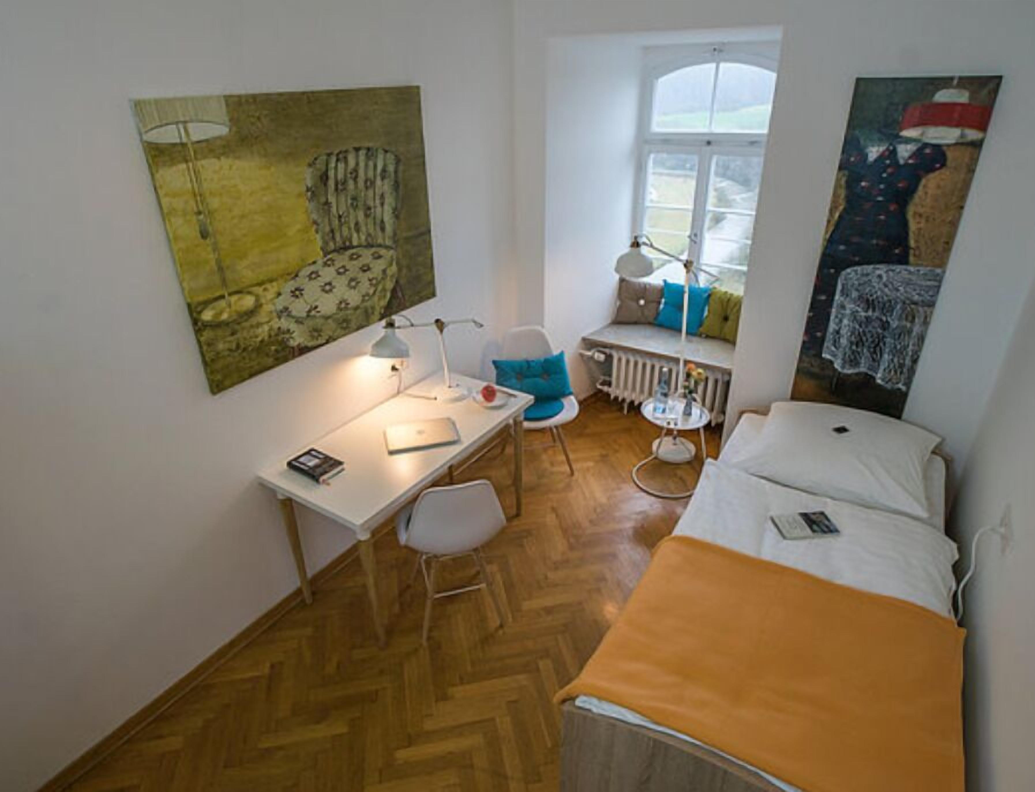 Une petite pièce bien éclairée, avec un plancher en bois, un bureau avec une chaise, un lit avec un matelas blanc, un radiateur et deux tableaux accrochés aux murs.