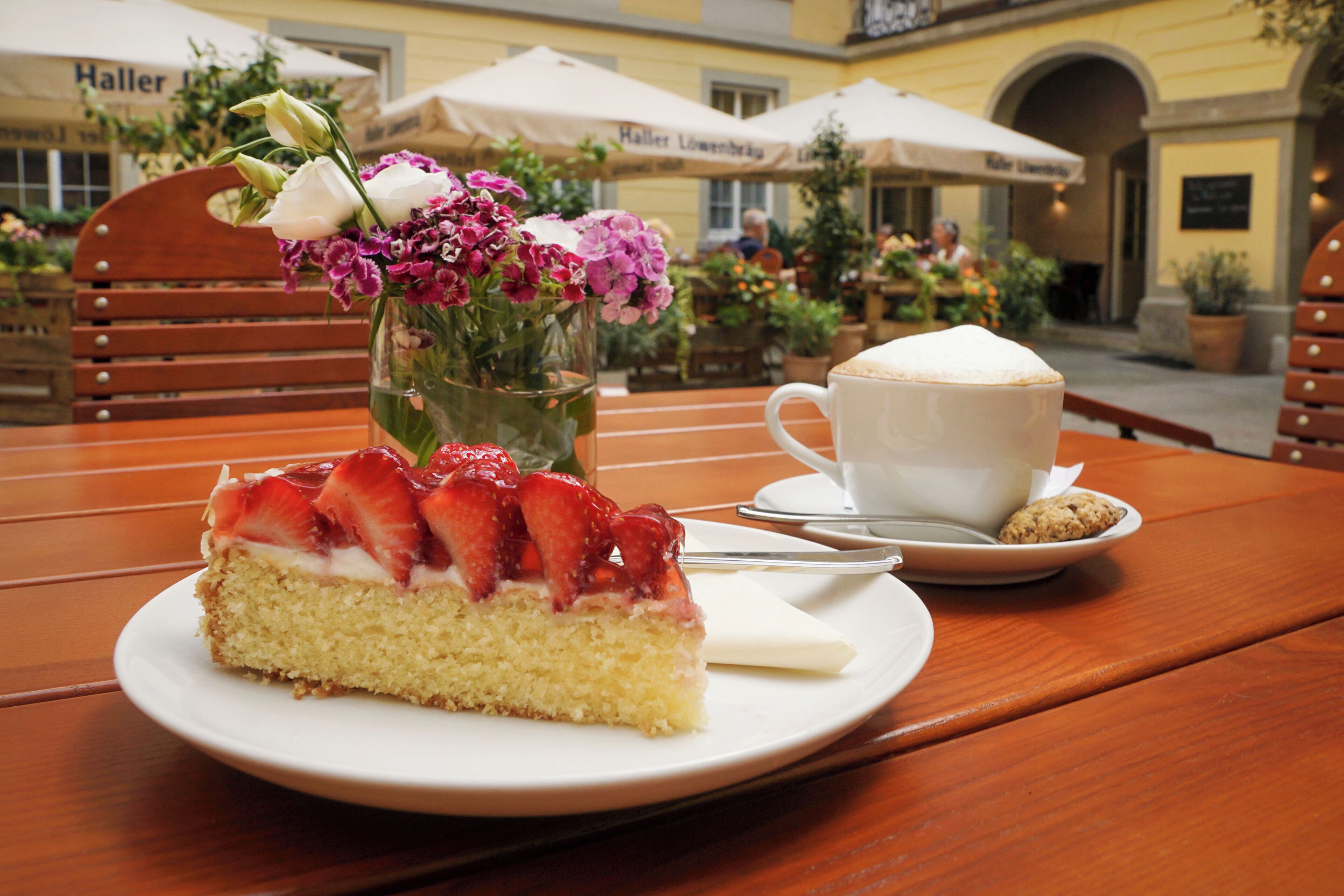 A slice of cake with strawberries on a white plate, a cup of coffee, and a vase of flowers on a wooden table.