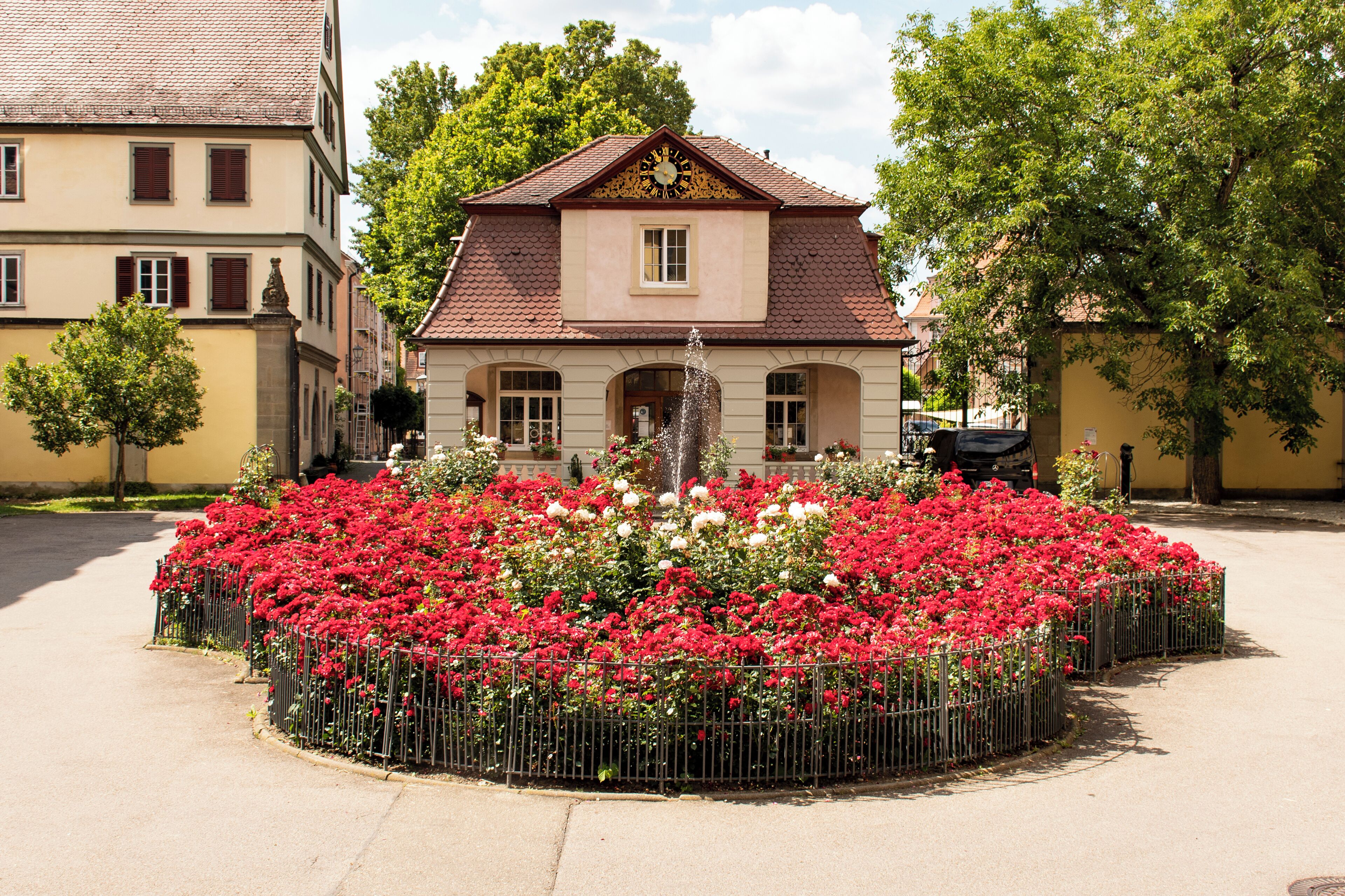 A building with a clock on the facade, surrounded by a circular flower bed with red and white flowers.