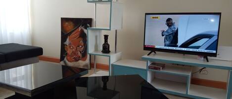 Un salón con una televisión de pantalla plana, una mesa de centro de vidrio, un cuadro y una botella decorativa sobre una estantería blanca.