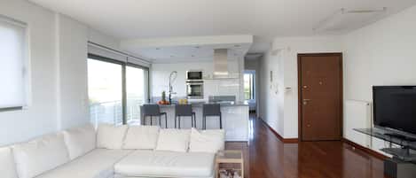 A modern living room with a white sofa, a glass coffee table with wine glasses and bottles, a TV on a stand, and a kitchen in the background.