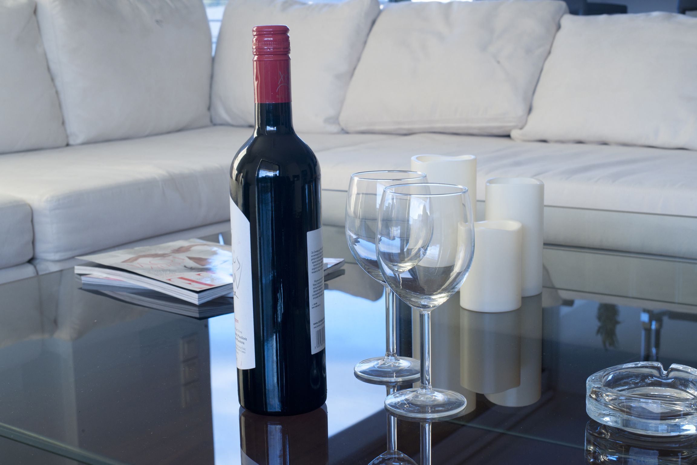 Une bouteille de vin, deux verres à vin et un cendrier sur une table en verre dans un salon avec un canapé blanc.