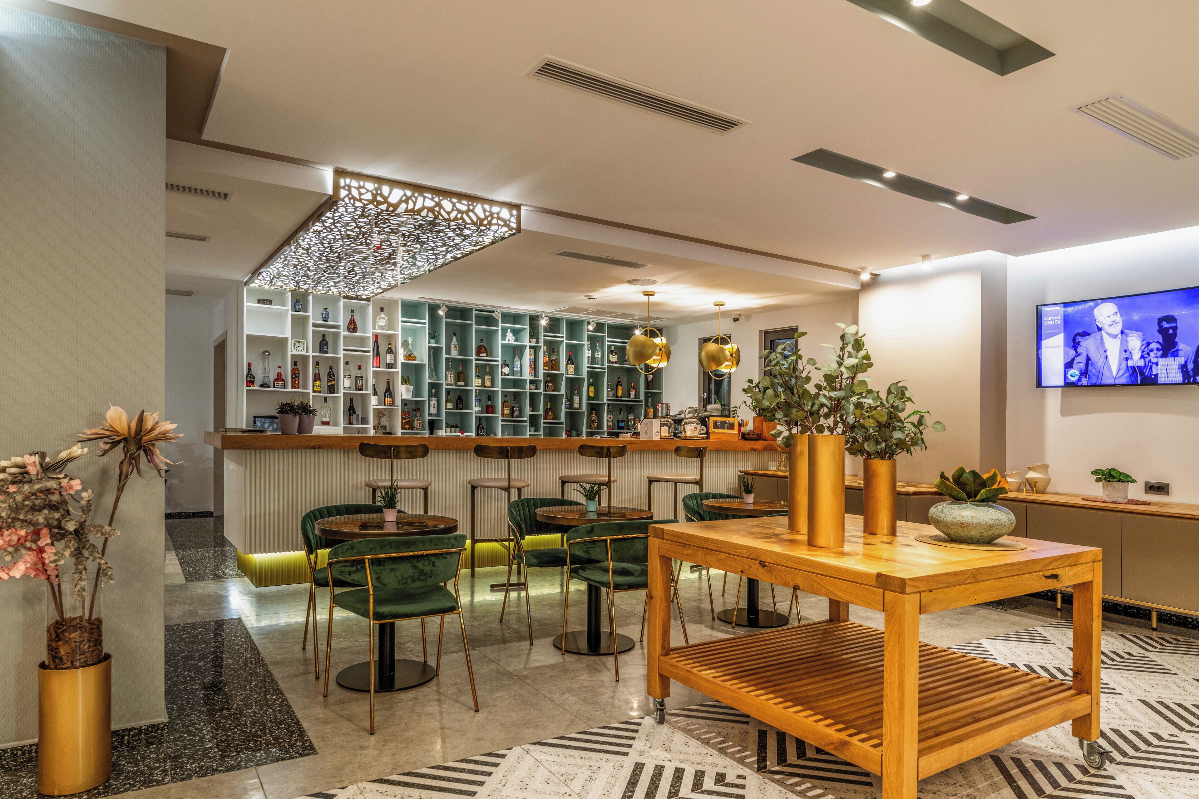 A modern bar area with green chairs, a wooden table, and a TV screen displaying a show.