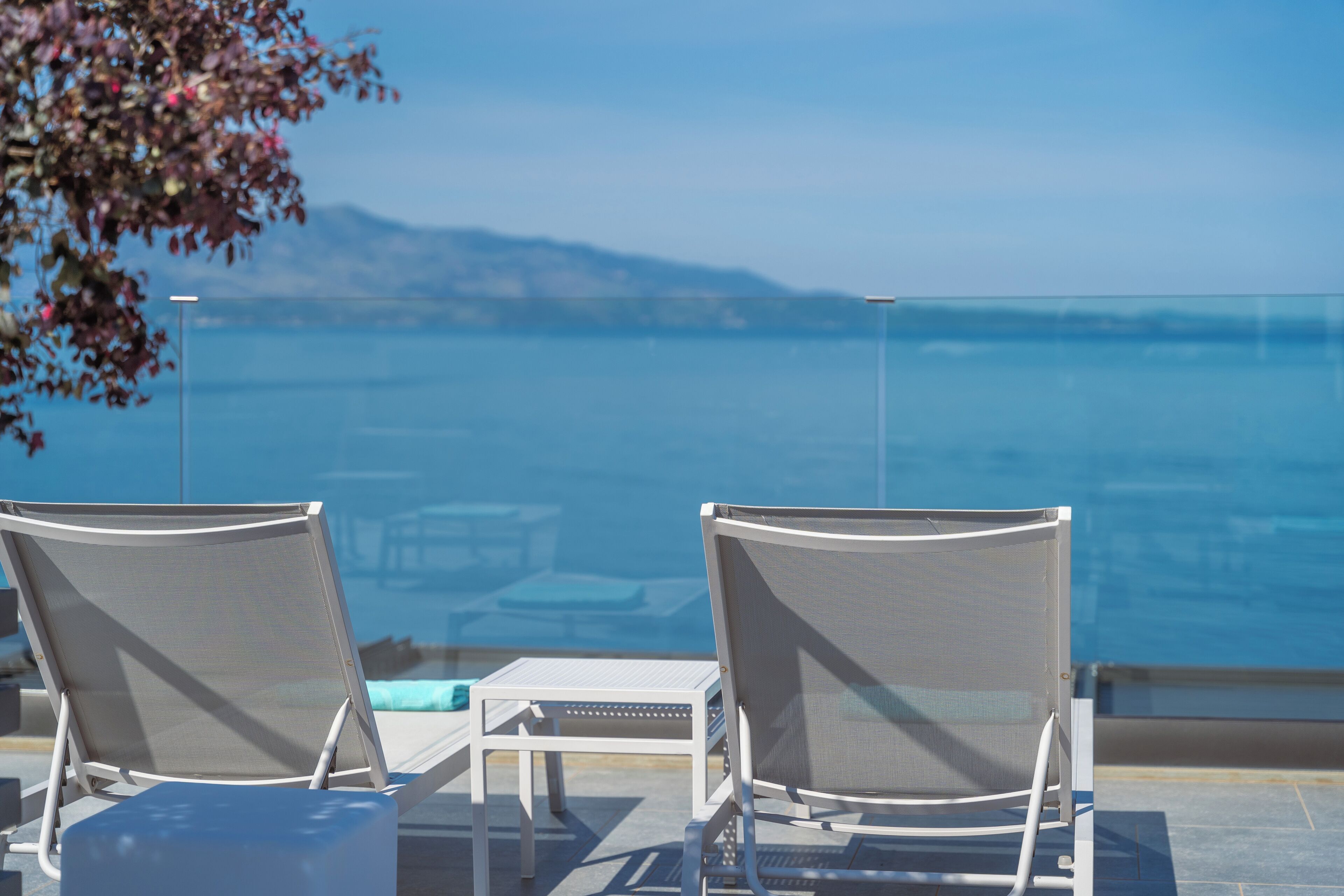 Two lounge chairs facing a pool with a clear view of the sea and distant mountains.