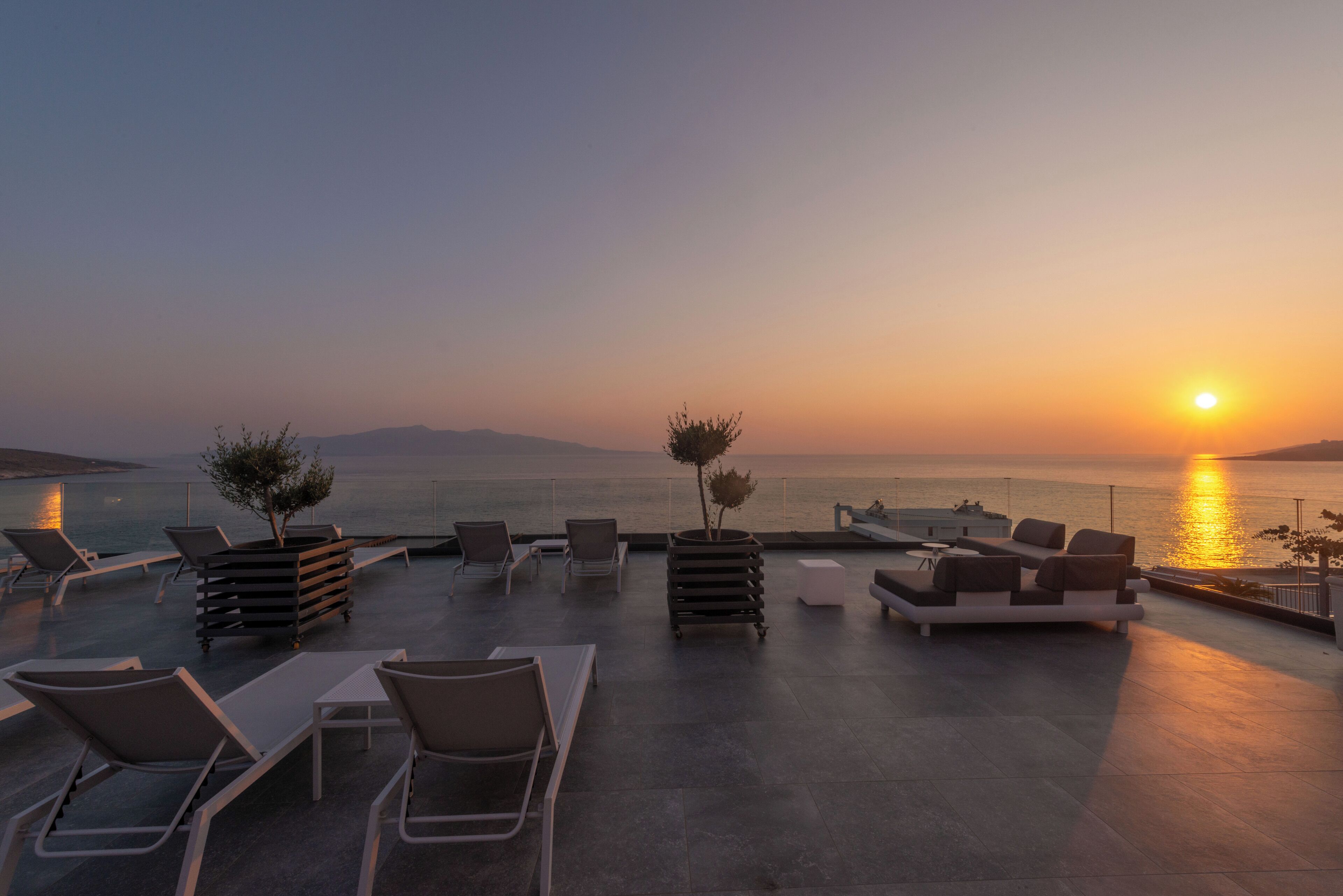 A rooftop terrace with lounge chairs and a small table, overlooking a sunset over the sea.