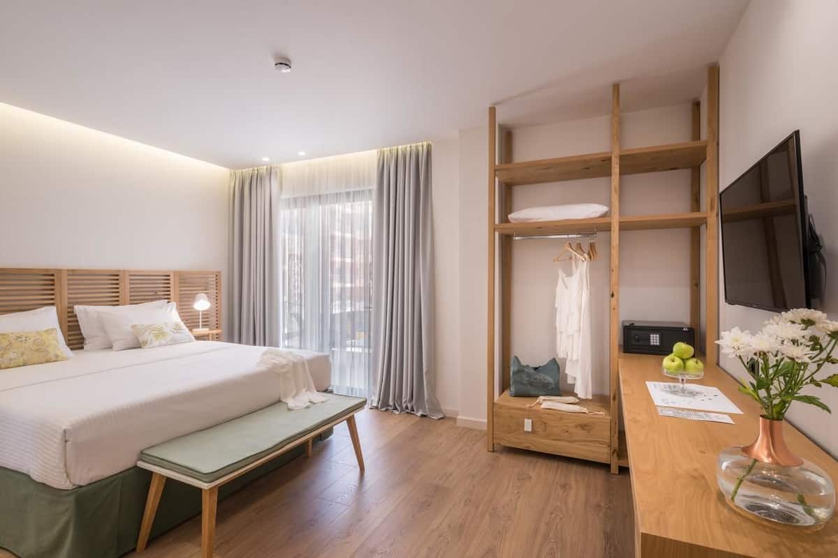 Hotellihuone, jossa on sänky, penkki, puinen työpöytä, televisio sekä hylly, jossa on vaatteita ja maljakko.