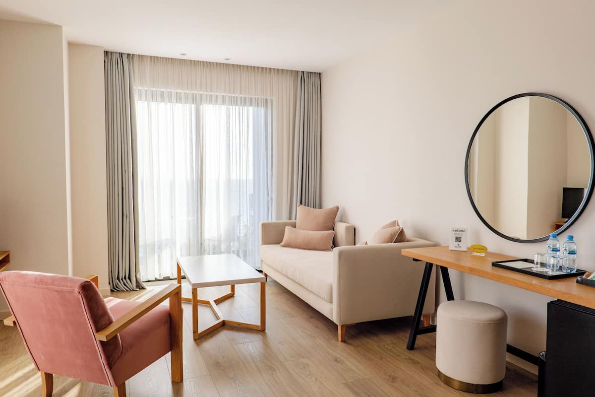 Hotellihuone, jossa on sohva, tuoli, pieni pöytä, pyöreä peili ja työpöytä, jonka päällä on tarjotinalusta.