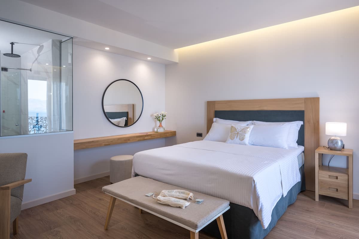 Moderni hotellihuone, jossa on suuri sänky, yöpöytä lampun kanssa, penkki pyyhkeillä, pyöreä peili ja lasiovesta avautuva näkymä kaupunkiin.
