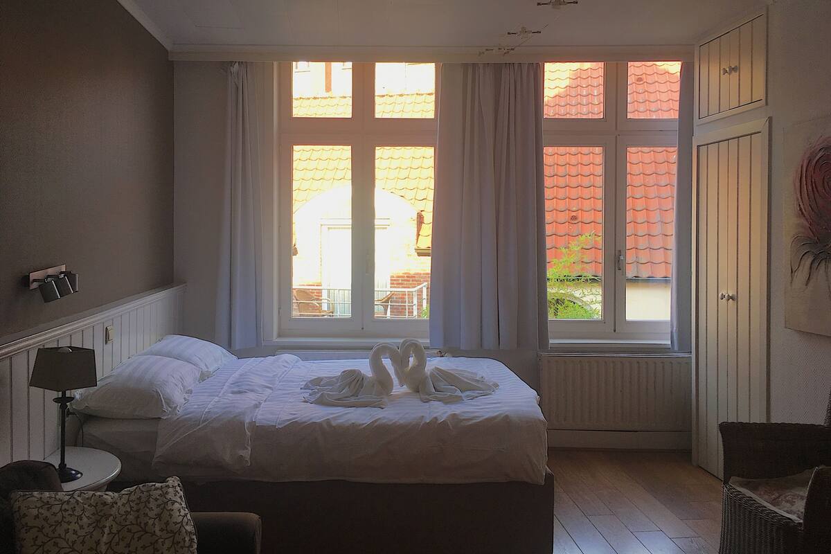 A hotel room with a bed, a window with curtains, a radiator, and a chair.