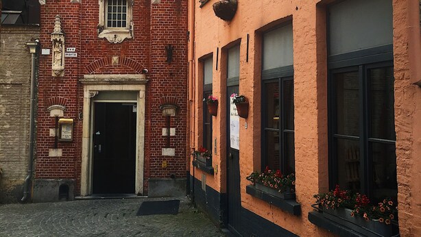 A narrow cobblestone street with a red brick building featuring a black door and a sign reading "Ave Maria". - Hotel Leopold (Bruges)