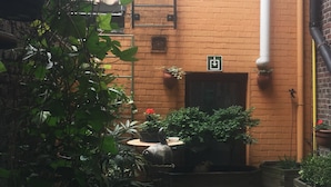 A courtyard with a brick wall, a window, and a door. There are potted plants and a small table with a chair. - Hotel Leopold (Bruges)