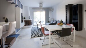 Una sala de estar moderna con un sofá blanco, una mesa de comedor con sillas, una mesa de centro y un televisor montado en la pared.