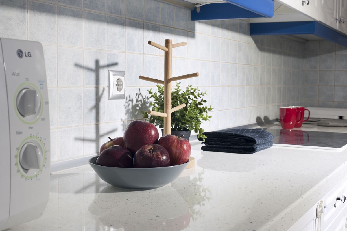 A kitchen with a white countertop, a bowl of apples, a wooden drying rack, a red mug, and a white appliance with green and white controls.