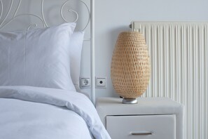 A neatly made bed with white linens, a white bedside table with a woven lamp, and a white radiator. - Glyfada, Modern Minimal Apartment (Glyfada)