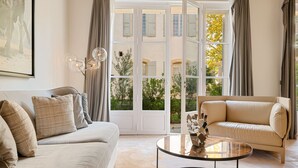 Una sala moderna con un sofá color crema, una mesa de centro de vidrio y un sillón color crema. Un amplio ventanal con cortinas ofrece vistas al exterior.
