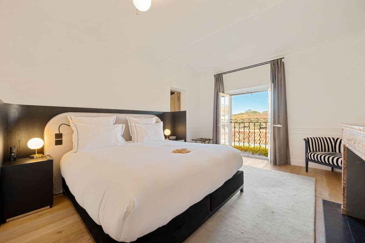 Ein modernes Schlafzimmer mit einem großen Bett, Nachttischlampen, Blick auf einen Weinberg durch eine Schiebetür und einem gestreiften Sessel am Kamin.
