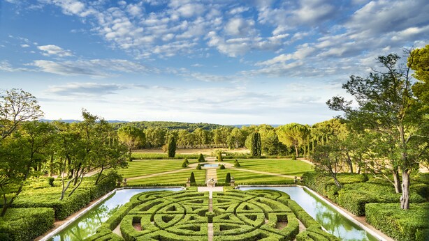 Un jardín formal con setos geométricos, una fuente central y cielo despejado.